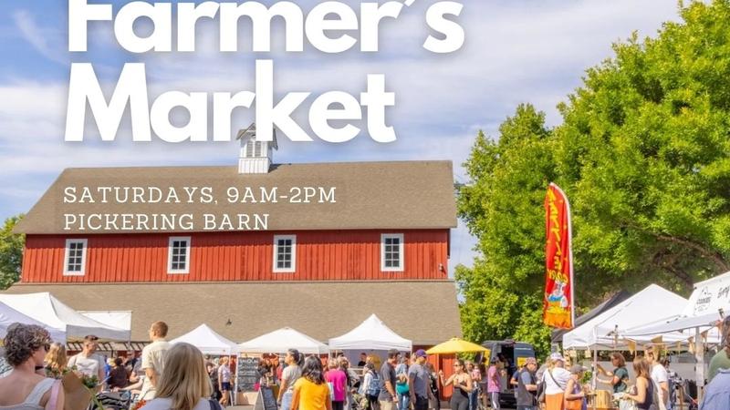 Shoppers and white tents at an outdoor farmers market, with produce crates and a red barn behind the crowd.