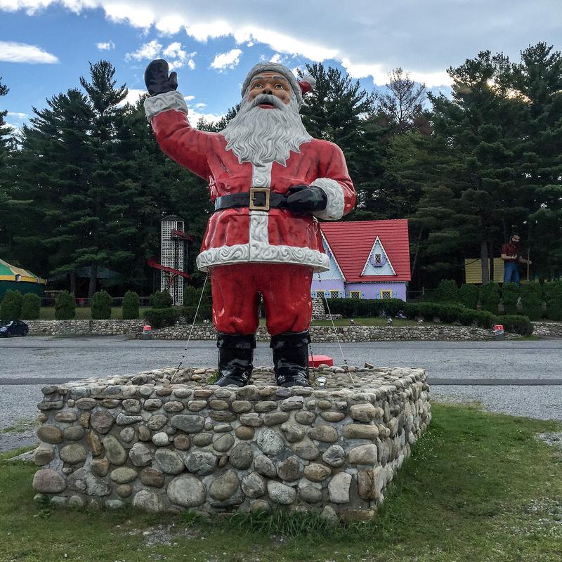 A seated Santa Claus statue in a red suit, outdoors by a fence.